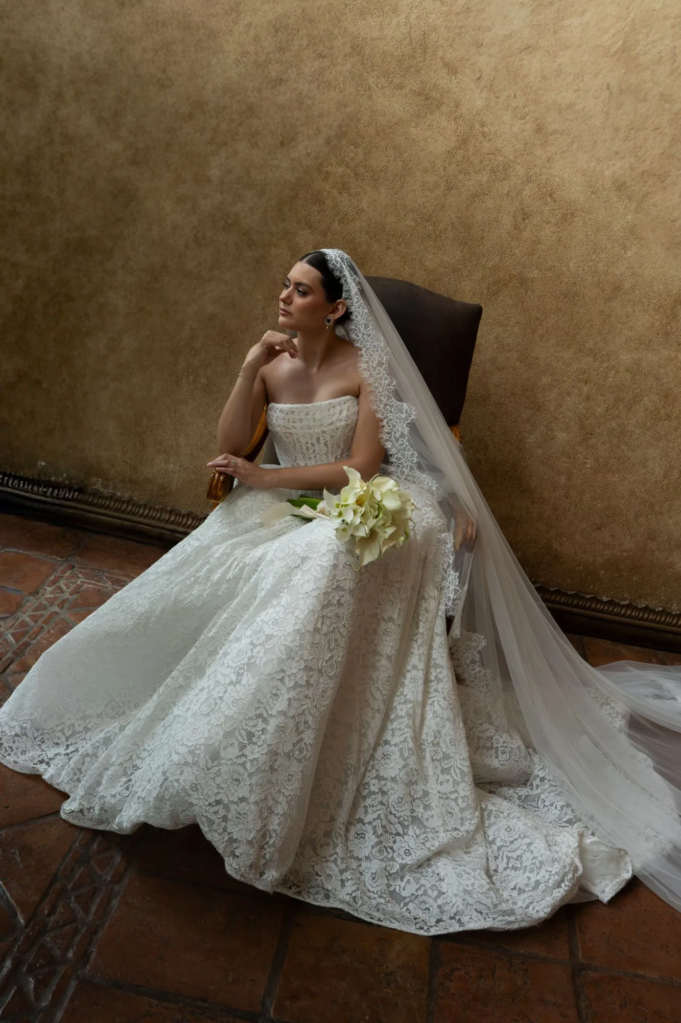A bride in a lace gown and long veil seated in a chair, holding calla lilies